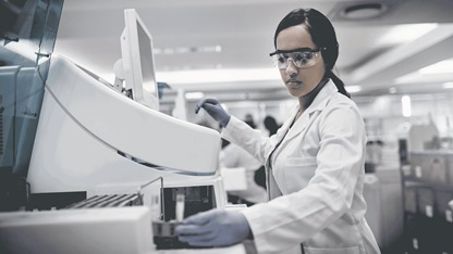 A lab technician in goggles, a white coat, and gloves uses a machine in a lab