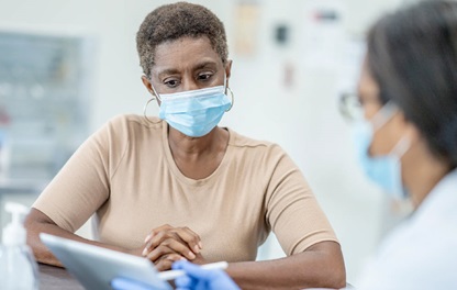 Photograph of a older african-american patient wearing a medical facemask looking at a tablet being held by a doctor in the foreground, out of focus