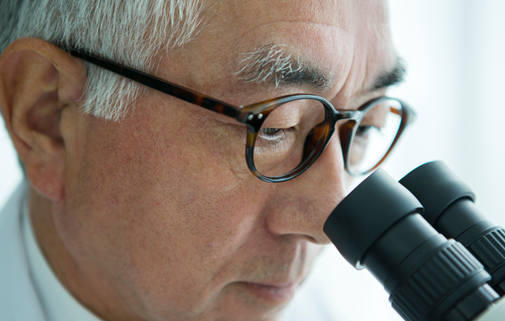Older man wearing glasses and a white lab coat looking intently through a microscope