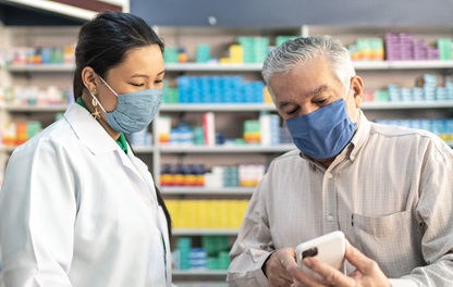 Man showing mobile device to woman wearing white coat, both wearing face masks, standing in front of shelves of medications