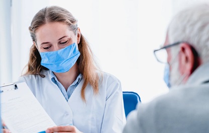 Photograph of a young female health care provider wearing a medical face mask consulting with an older male patient wearing a mask
