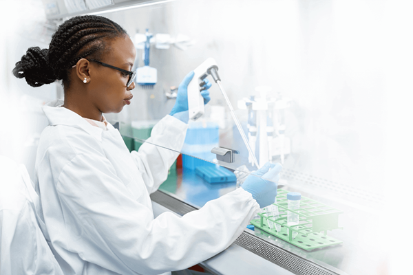A photograph of a female technician, wearing hair braids, glasses, a white lab coat, and blue latex gloves, focusing intently on her work in a biopharmaceutical lab