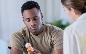 A photograph of a young african-amercan man reading the label of a medication bottle, while a health care provider sits across from him, out of focus, in the foreground