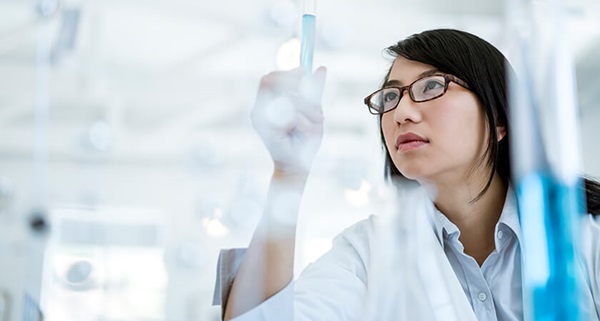 Photograph of a woman in lab clothes and a collared shirt examining a test tube filled with blue liquid